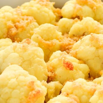 Close-up of baked cauliflower florets topped with golden, crispy Parmesan and pancetta crumbles in a white rectangular baking dish, with a blue striped kitchen towel in the background. No text on image.