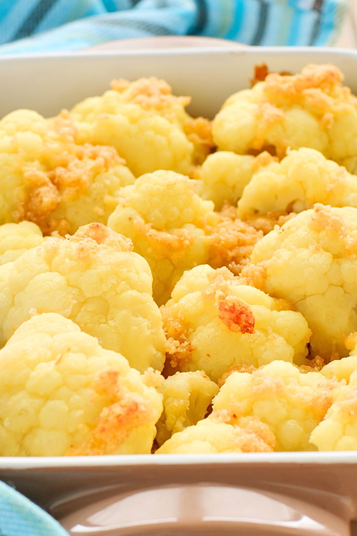 Close-up of baked cauliflower florets topped with golden, crispy Parmesan and pancetta crumbles in a white rectangular baking dish, with a blue striped kitchen towel in the background. No text on image.