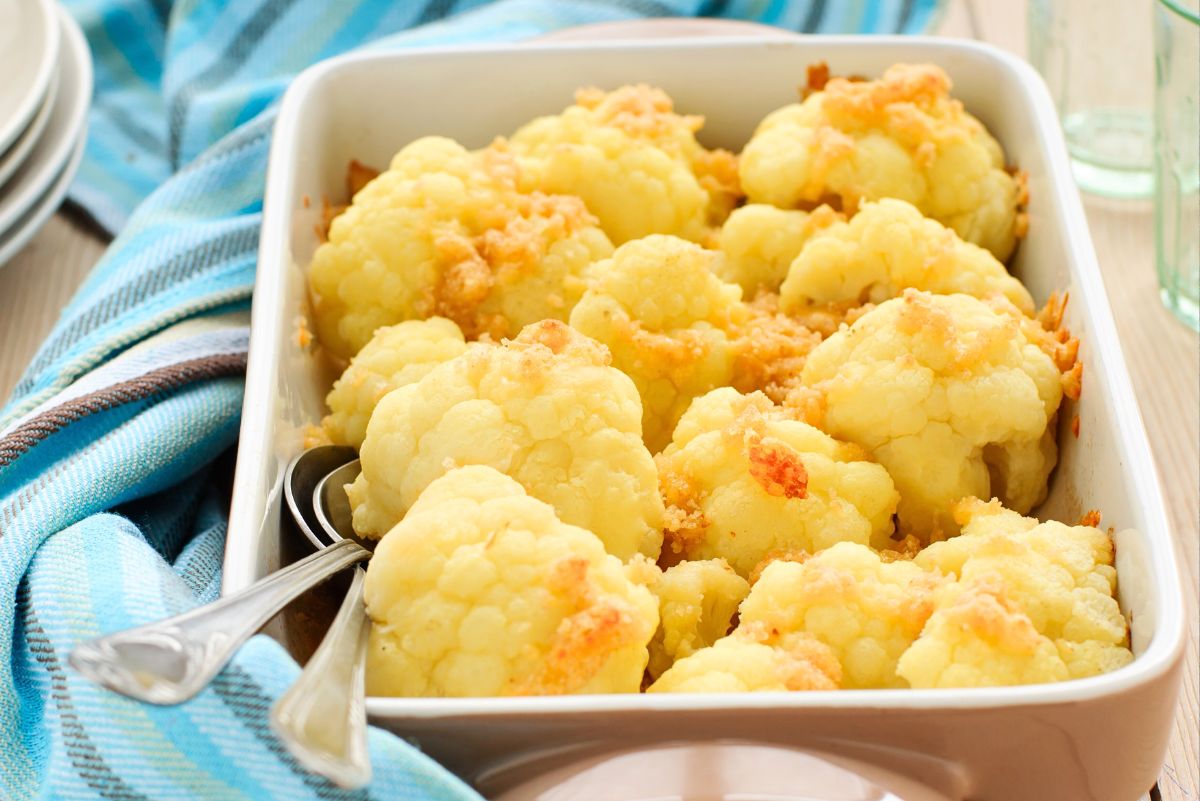 A white rectangular baking dish filled with baked cauliflower florets topped with golden, crispy Parmesan and pancetta, with two silver serving spoons resting inside the dish. A blue striped kitchen towel, a stack of white plates, and a glass are visible in the background on a light wooden surface. No text on image.