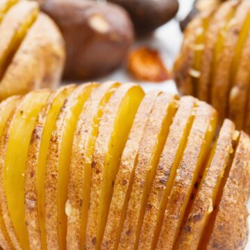 Close-up of baked Hasselback potato sliced into thin accordion layers, roasted until golden brown with crispy edges and tender yellow center. Skin-on potato with visible seasoning on a white plate, mushrooms blurred in the background. Crispy baked Hasselback potatoes with Romano cheese and roasted garlic.