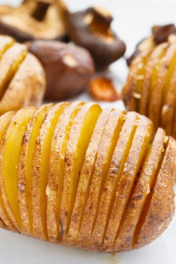 Close-up of baked Hasselback potato sliced into thin accordion layers, roasted until golden brown with crispy edges and tender yellow center. Skin-on potato with visible seasoning on a white plate, mushrooms blurred in the background. Crispy baked Hasselback potatoes with Romano cheese and roasted garlic.