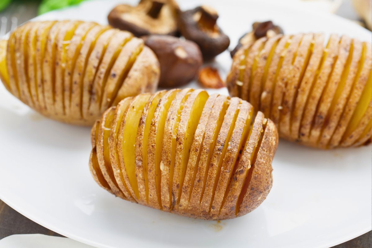 Three baked Hasselback potatoes on a white plate, thinly sliced into even accordion layers and roasted until golden brown with crispy edges and tender centers. Skin-on potatoes with light seasoning, mushrooms in the background. Crispy baked Hasselback potatoes with Romano cheese, perfect side dish.