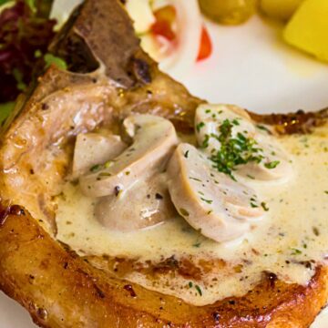 Baked pork chop with mushroom gravy on a white plate, featuring a bone in pork chop topped with creamy mushroom sauce and sliced mushrooms, sprinkled with parsley. Side salad and boiled potatoes in the background, highlighting a high protein dinner with rich, savory flavors.
