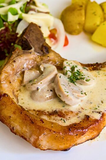 Baked pork chop with mushroom gravy on a white plate, featuring a bone in pork chop topped with creamy mushroom sauce and sliced mushrooms, sprinkled with parsley. Side salad and boiled potatoes in the background, highlighting a high protein dinner with rich, savory flavors.
