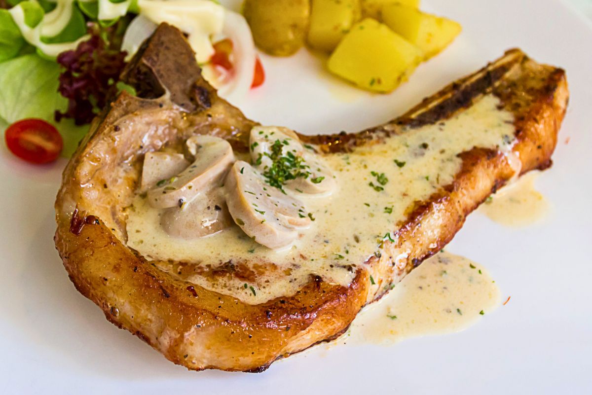 Baked pork chop with mushroom gravy on a white plate, featuring a bone in pork chop topped with creamy mushroom sauce and sliced mushrooms, garnished with parsley. Boiled potatoes and a small side salad with cherry tomatoes in the background, showcasing a hearty high protein dinner.