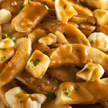 Close up of Canadian poutine on a white plate with crispy golden French fries topped with fresh cheese curds and rich brown gravy, garnished with chopped parsley. The gravy melts slightly into the cheese curds and coats the fries for a classic homemade comfort food dish.