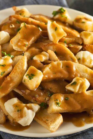 Close up of Canadian poutine on a white plate with crispy golden French fries topped with fresh cheese curds and rich brown gravy, garnished with chopped parsley. The gravy melts slightly into the cheese curds and coats the fries for a classic homemade comfort food dish.