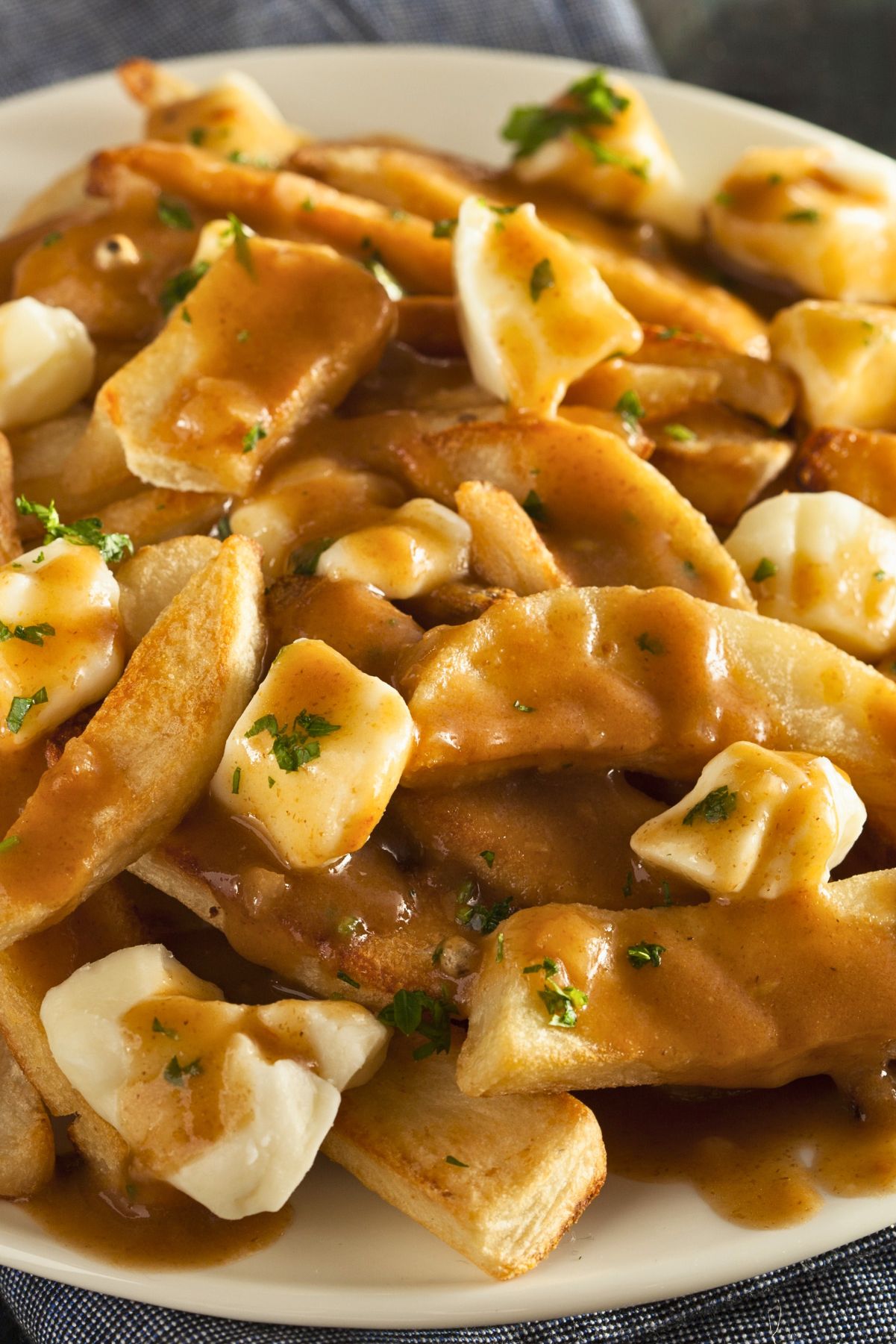 Close up of Canadian poutine on a white plate with crispy golden French fries topped with fresh cheese curds and rich brown gravy, garnished with chopped parsley. The gravy melts slightly into the cheese curds and coats the fries for a classic homemade comfort food dish.