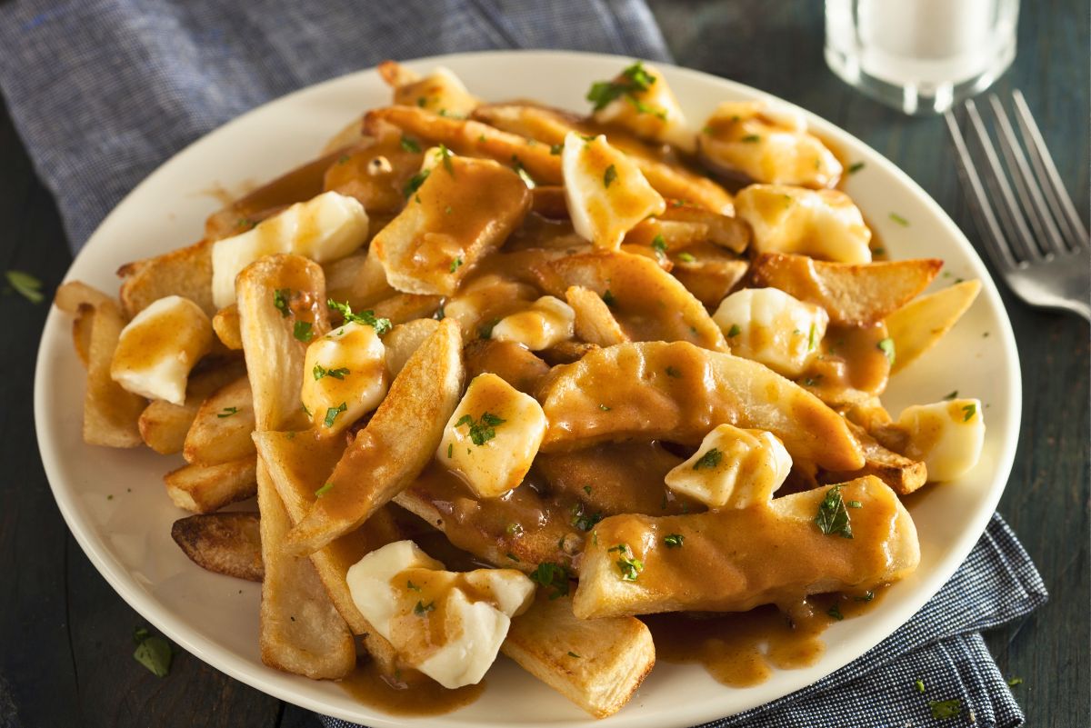 White plate of Canadian poutine with thick cut crispy French fries topped with fresh white cheese curds and rich brown gravy, sprinkled with chopped parsley. Served on a rustic wooden table with a fork and napkin in the background, classic homemade comfort food.