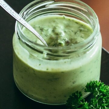 Close-up of creamy green goddess dressing with avocado in a small glass jar, a silver spoon inside. Fresh parsley sprigs rest beside the jar on a dark countertop. Thick, smooth homemade avocado herb dressing with visible flecks of dill and herbs, perfect for salads or as a dip.