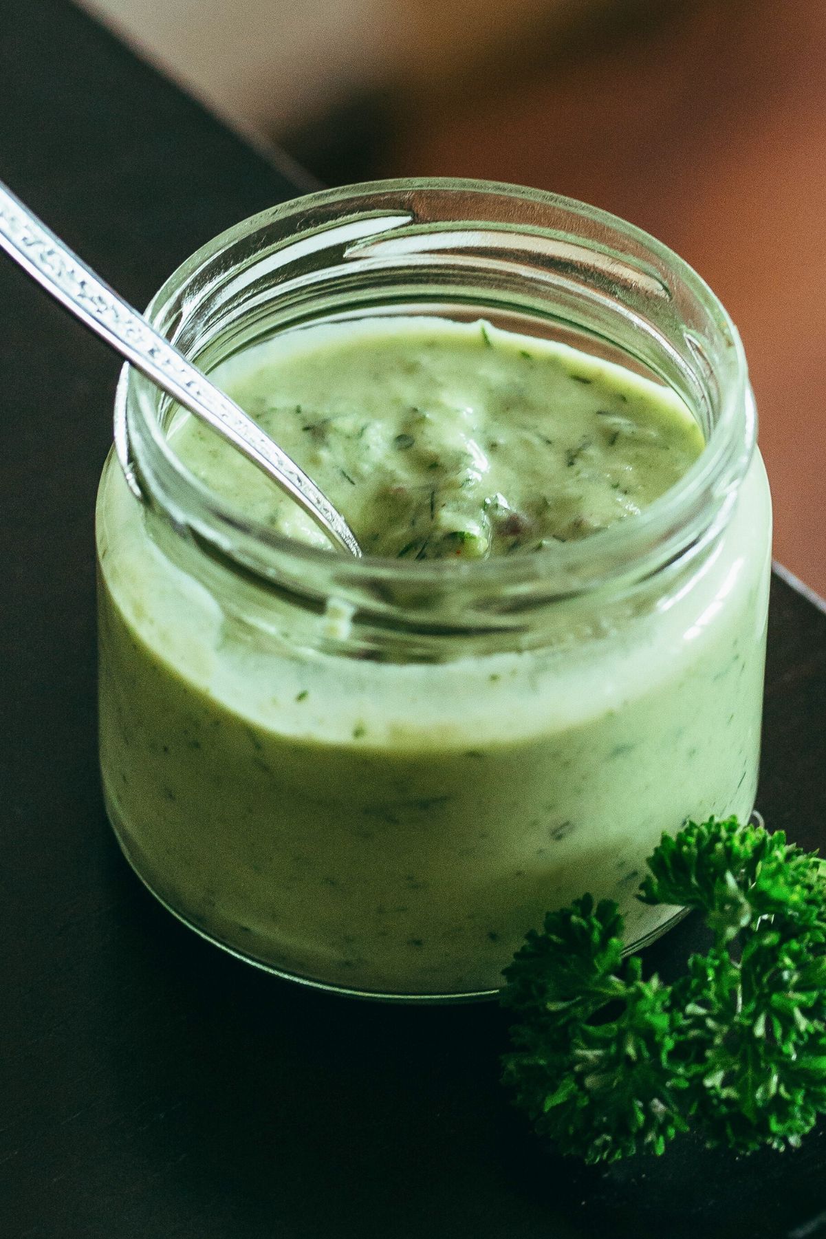 Close-up of creamy green goddess dressing with avocado in a small glass jar, a silver spoon inside. Fresh parsley sprigs rest beside the jar on a dark countertop. Thick, smooth homemade avocado herb dressing with visible flecks of dill and herbs, perfect for salads or as a dip.