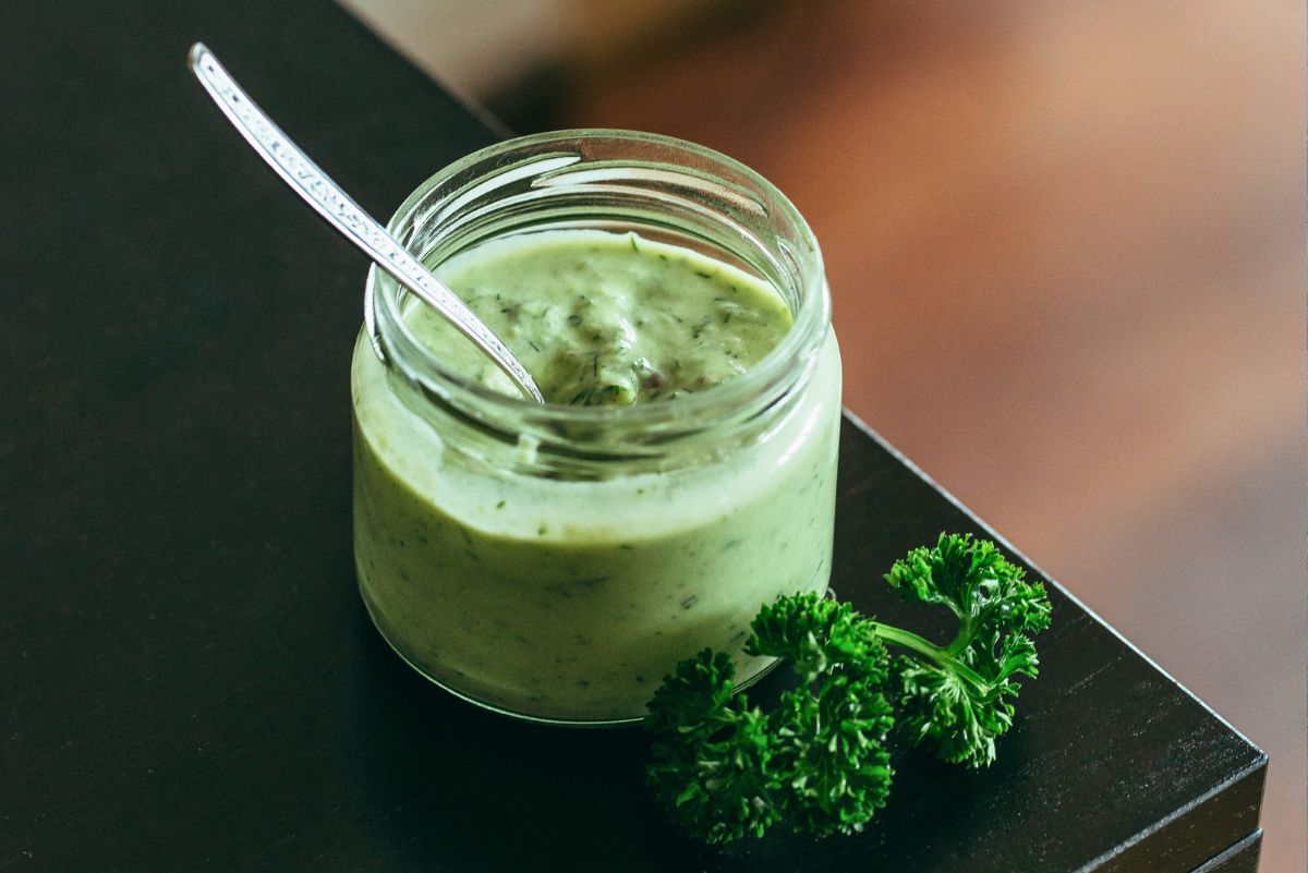 Small glass jar filled with creamy green goddess dressing made with avocado, a silver spoon resting inside. Fresh parsley sprigs sit beside the jar on a dark countertop. Thick, smooth homemade avocado herb salad dressing with visible flecks of dill and green herbs.