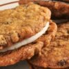 Close up of copycat Little Debbie oatmeal cream pies stacked on a plate, chewy oatmeal cookies with visible oats and raisins sandwiched around thick white cream filling. Soft textured homemade sandwich cookies with lightly crisp edges and creamy center.
