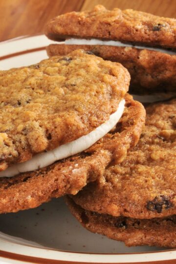 Close up of copycat Little Debbie oatmeal cream pies stacked on a plate, chewy oatmeal cookies with visible oats and raisins sandwiched around thick white cream filling. Soft textured homemade sandwich cookies with lightly crisp edges and creamy center.