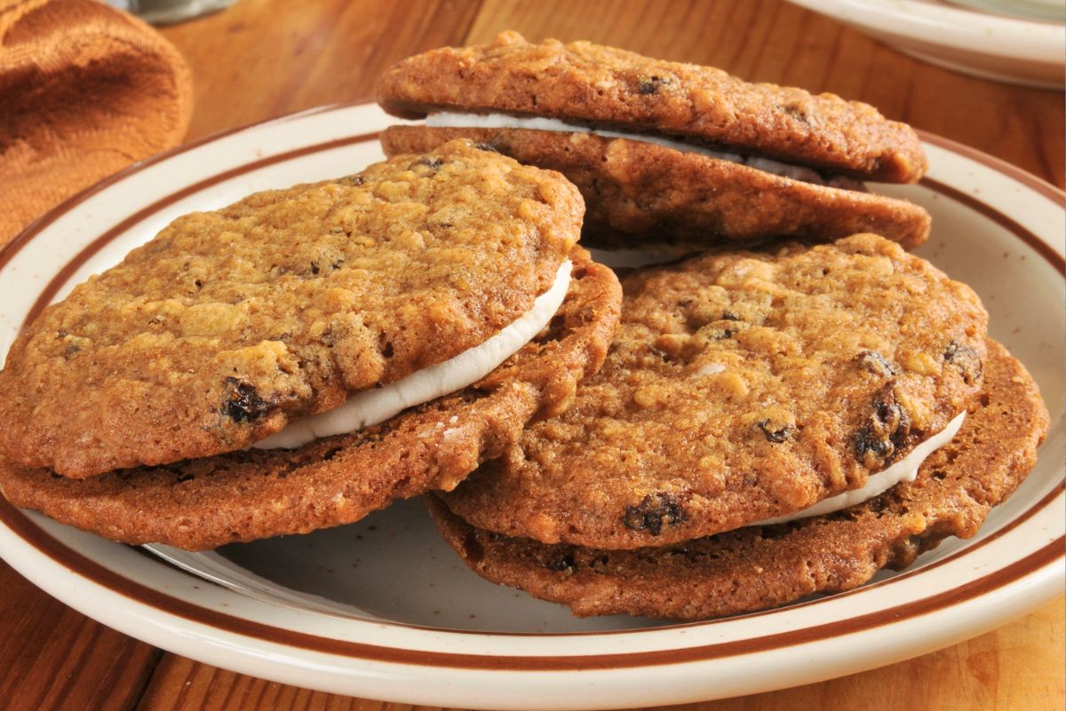 Plate of copycat Little Debbie oatmeal cream pies with soft, chewy oatmeal cookies filled with thick white cream. Visible oats and raisins in golden brown sandwich cookies stacked on a rustic plate, homemade dessert treat on a wooden table.