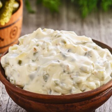 Wooden bowl filled with creamy homemade tartar sauce, thick and chunky with visible pickle relish and onion pieces. Close up of copycat McDonald&rsquo;s tartar sauce on a rustic wooden table, with pickles and fresh herbs blurred in the background.