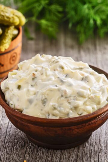 Wooden bowl filled with creamy homemade tartar sauce, thick and chunky with visible pickle relish and onion pieces. Close up of copycat McDonald’s tartar sauce on a rustic wooden table, with pickles and fresh herbs blurred in the background.
