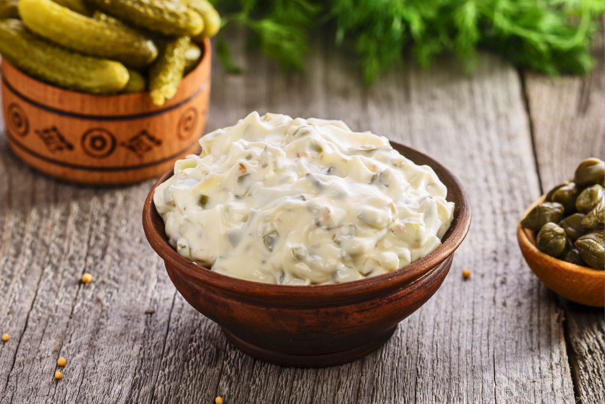 Wooden bowl of creamy copycat McDonald&rsquo;s tartar sauce with visible pickle relish and onion pieces, thick and smooth texture. Set on a rustic wooden table with pickles and capers in small bowls and fresh dill in the background, homemade condiment for fish and seafood.