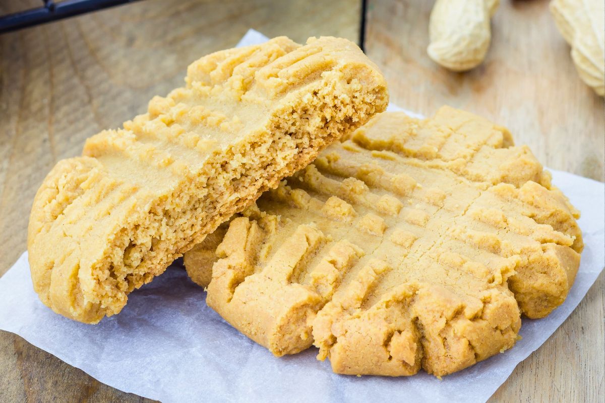Thick copycat Mrs. Fields peanut butter cookies with classic fork crisscross pattern, soft and crumbly texture. One cookie is split open to show the tender peanut butter interior, resting on parchment paper on a wooden surface with a cooling rack and whole peanuts in the background.