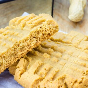 Close up of copycat Mrs. Fields peanut butter cookies with classic fork crisscross marks, soft and thick with a tender crumb. One cookie is broken in half showing the moist interior, resting on parchment paper with a cooling rack and whole peanut in the background.