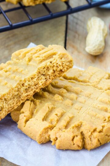 Close up of copycat Mrs. Fields peanut butter cookies with classic fork crisscross marks, soft and thick with a tender crumb. One cookie is broken in half showing the moist interior, resting on parchment paper with a cooling rack and whole peanut in the background.
