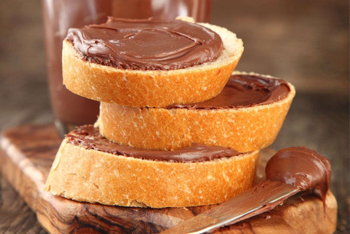 Stack of thick bread slices spread with glossy chocolate hazelnut spread on a rustic wooden board. Close up of creamy homemade chocolate hazelnut spread with a knife coated in chocolate and a glass of chocolate drink in the background.