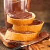 Slices of toasted bread stacked with thick chocolate hazelnut spread, glossy and smooth, on a rustic wooden board. Knife with chocolate spread and a glass of chocolate hazelnut drink in the background, homemade copycat Nutella style chocolate spread.