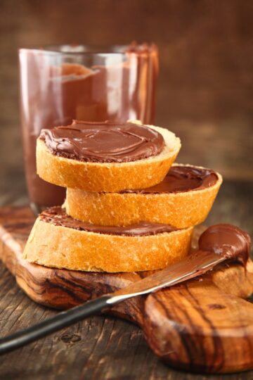 Slices of toasted bread stacked with thick chocolate hazelnut spread, glossy and smooth, on a rustic wooden board. Knife with chocolate spread and a glass of chocolate hazelnut drink in the background, homemade copycat Nutella style chocolate spread.