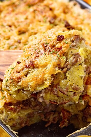 Close-up of ground beef and potato bake layered with thin sliced potatoes, seasoned beef, and melted cheddar cheese. Golden brown cheesy crust with crispy edges, a generous square lifted from a black baking dish on a wooden spatula. Hearty comfort food casserole dinner.