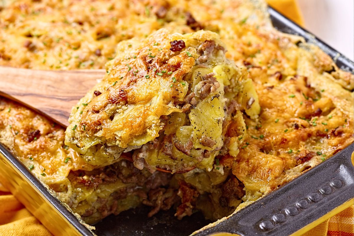 Ground beef and potato bake with layers of thin sliced potatoes, seasoned beef, and melted cheddar cheese. Golden brown cheesy top with crispy edges, a thick serving lifted from a black casserole dish on a wooden spatula. Hearty comfort food dinner casserole garnished with chopped herbs.