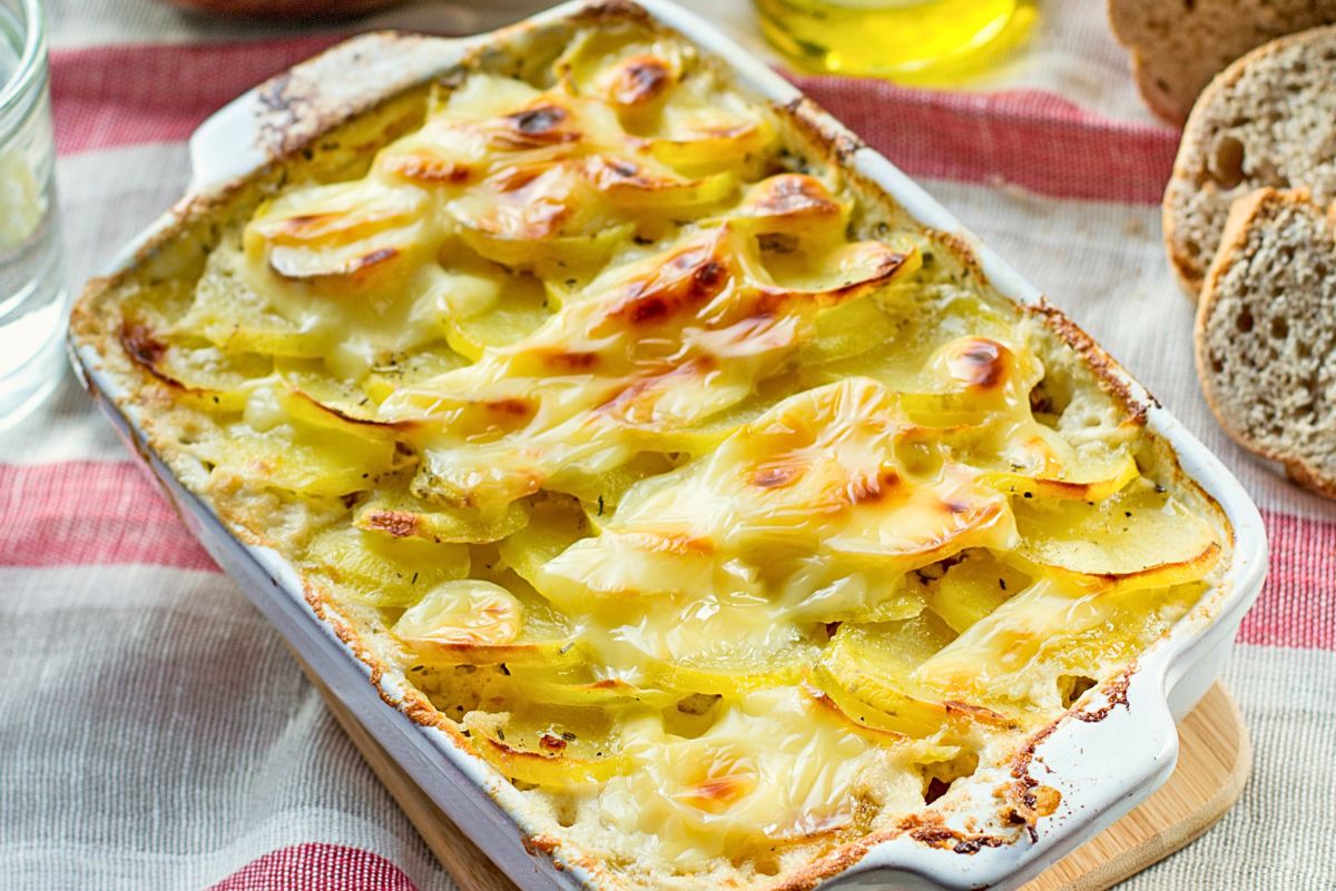 Garlic potato bake with bacon and cabbage in a white ceramic casserole dish, layered sliced potatoes in a creamy sauce topped with melted, lightly browned cheese. Golden, crispy edges and bubbling sauce visible around the sides, served on a wooden board with sliced bread in the background.