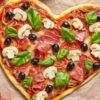 Heart shaped pizza on parchment paper topped with prosciutto, sliced mushrooms, black olives, tomato slices, melted cheese, and fresh basil leaves. Golden crust with red sauce visible underneath, styled on a gray stone surface with whole tomatoes nearby.