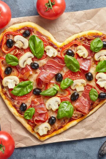 Heart shaped pizza on parchment paper topped with prosciutto, sliced mushrooms, black olives, tomato slices, melted cheese, and fresh basil leaves. Golden crust with red sauce visible underneath, styled on a gray stone surface with whole tomatoes nearby.