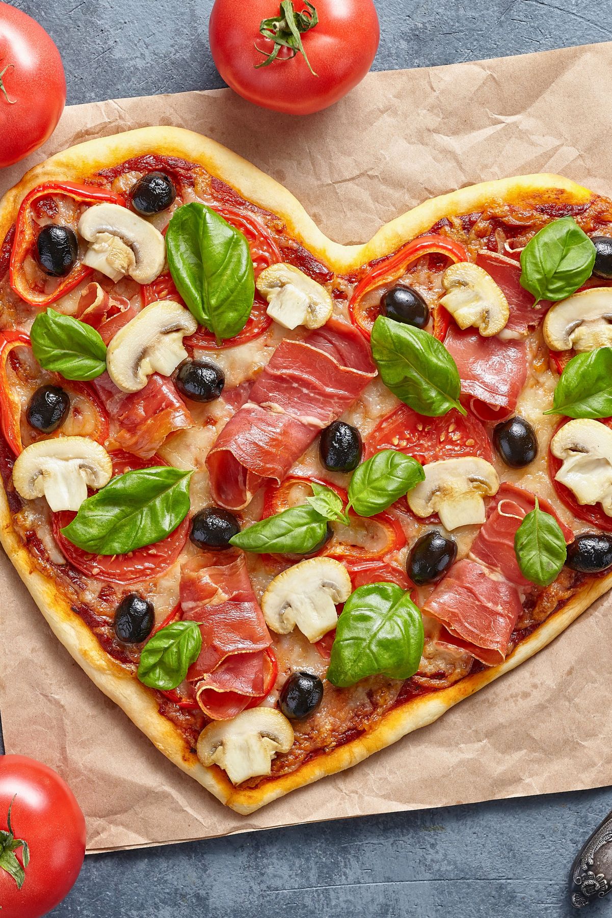 Heart shaped pizza on parchment paper topped with prosciutto, sliced mushrooms, black olives, tomato slices, melted cheese, and fresh basil leaves. Golden crust with red sauce visible underneath, styled on a gray stone surface with whole tomatoes nearby.