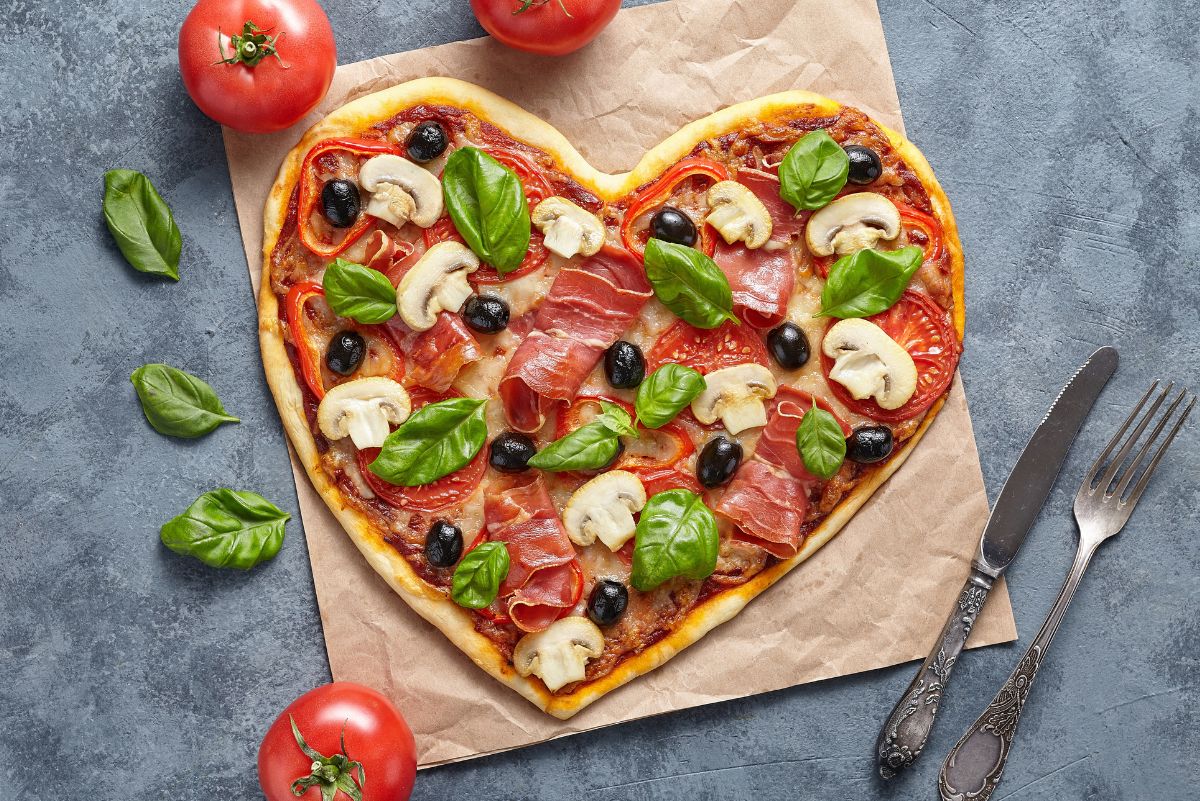 Heart shaped pizza on parchment paper topped with prosciutto, sliced mushrooms, black olives, tomato slices, melted cheese, and fresh basil leaves. Golden crust with visible red sauce, styled on a gray stone surface with whole tomatoes, basil leaves, and a fork and knife nearby.