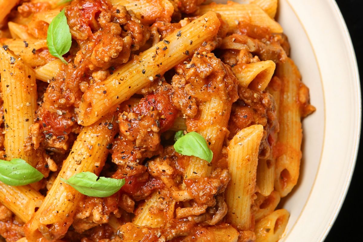 Bowl of Italian beef ragu with penne pasta coated in rich tomato meat sauce, topped with cracked black pepper and fresh basil leaves. Tender ground beef and tomatoes cling to ridged penne for a hearty high protein Italian pasta dinner.