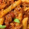Close-up of Italian beef ragu with penne pasta coated in rich tomato sauce and tender ground beef, sprinkled with cracked black pepper and fresh basil leaves. Hearty high protein pasta dinner with thick, savory meat sauce over ridged penne.