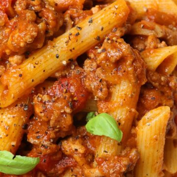 Close-up of Italian beef ragu with penne pasta coated in rich tomato sauce and tender ground beef, sprinkled with cracked black pepper and fresh basil leaves. Hearty high protein pasta dinner with thick, savory meat sauce over ridged penne.