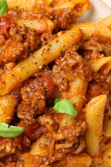 Close-up of Italian beef ragu with penne pasta coated in rich tomato sauce and tender ground beef, sprinkled with cracked black pepper and fresh basil leaves. Hearty high protein pasta dinner with thick, savory meat sauce over ridged penne.