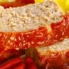 Close-up of sliced high protein baked meatloaf with thick honey ketchup topping dripping down the sides. Juicy, tender meatloaf on a white plate with glossy tomato glaze, red chili peppers in front and a yellow napkin blurred in the background. Homemade protein-packed meatloaf with sweet and tangy ketchup glaze.
