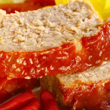 Close-up of sliced high protein baked meatloaf with thick honey ketchup topping dripping down the sides. Juicy, tender meatloaf on a white plate with glossy tomato glaze, red chili peppers in front and a yellow napkin blurred in the background. Homemade protein-packed meatloaf with sweet and tangy ketchup glaze.