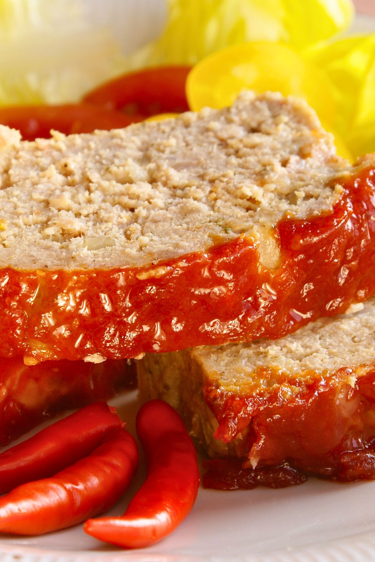 Close-up of sliced high protein baked meatloaf with thick honey ketchup topping dripping down the sides. Juicy, tender meatloaf on a white plate with glossy tomato glaze, red chili peppers in front and a yellow napkin blurred in the background. Homemade protein-packed meatloaf with sweet and tangy ketchup glaze.