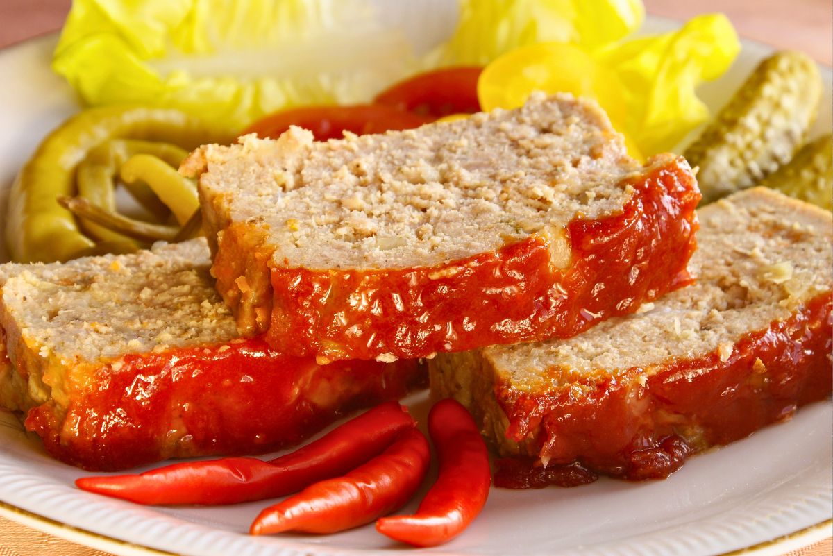 Three slices of high protein baked meatloaf stacked on a white plate, coated in thick honey ketchup topping. Glossy tomato glaze drips down the sides. Red chili peppers in front, with pickles, pepper rings, and lettuce blurred in the background. Juicy homemade meatloaf with sweet and tangy ketchup glaze.