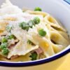 Close-up of ham and cheese pasta casserole in a white bowl with blue rim, made with bowtie pasta, diced ham, green peas, and creamy cheese sauce, topped with grated Parmesan. Comforting high protein pasta dinner with tender noodles and savory ham.