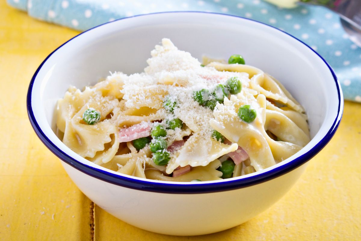 White bowl with blue rim filled with ham and cheese pasta casserole made with bowtie pasta, diced ham, green peas, and creamy cheese sauce, topped with grated Parmesan. Set on a yellow table with a blue polka dot napkin in the background. High protein comfort food pasta dinner recipe.