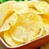Homemade potato chips from scratch in a small square bowl, thin sliced golden crispy chips with lightly browned edges. Bowl sits on a rustic wooden table with fresh parsley and whole potatoes in the background. Crunchy, classic snack made from fresh sliced potatoes.