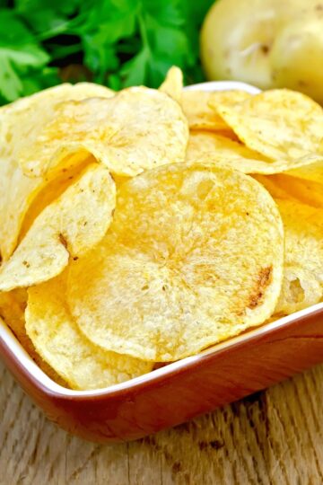 Homemade potato chips from scratch in a small square bowl, thin sliced golden crispy chips with lightly browned edges. Bowl sits on a rustic wooden table with fresh parsley and whole potatoes in the background. Crunchy, classic snack made from fresh sliced potatoes.