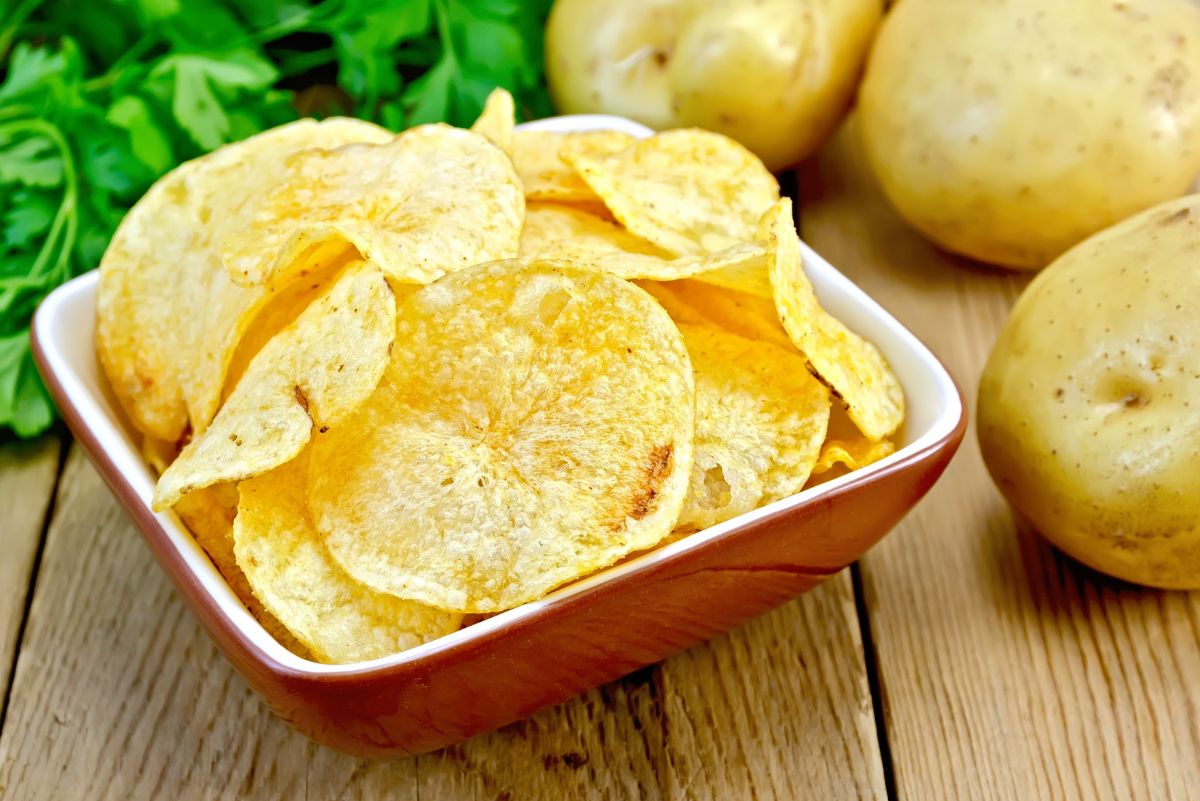 Homemade potato chips from scratch in a small square bowl, thin sliced golden crispy chips with lightly browned edges. Bowl sits on a rustic wooden table with fresh parsley and whole raw potatoes in the background, highlighting a classic crunchy snack made from fresh potatoes.