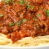Close-up of Italian sausage pasta dinner with spaghetti topped in chunky tomato sauce, browned Italian sausage, diced tomatoes, and fresh chopped parsley. Rich red marinara over tender pasta in a white bowl. Hearty high protein pasta recipe, classic comfort food family dinner.