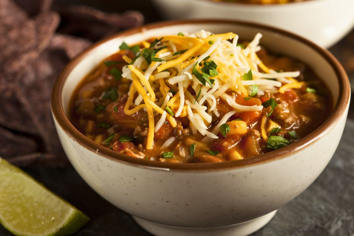 Bowl of Southwest beef and corn chili topped with shredded cheddar and Monterey Jack cheese and fresh chopped green onions. Thick tomato-based chili with ground beef, corn, and beans in a speckled ceramic bowl. Lime wedge and blue corn tortilla chips on the side. Hearty high protein one pot dinner recipe.